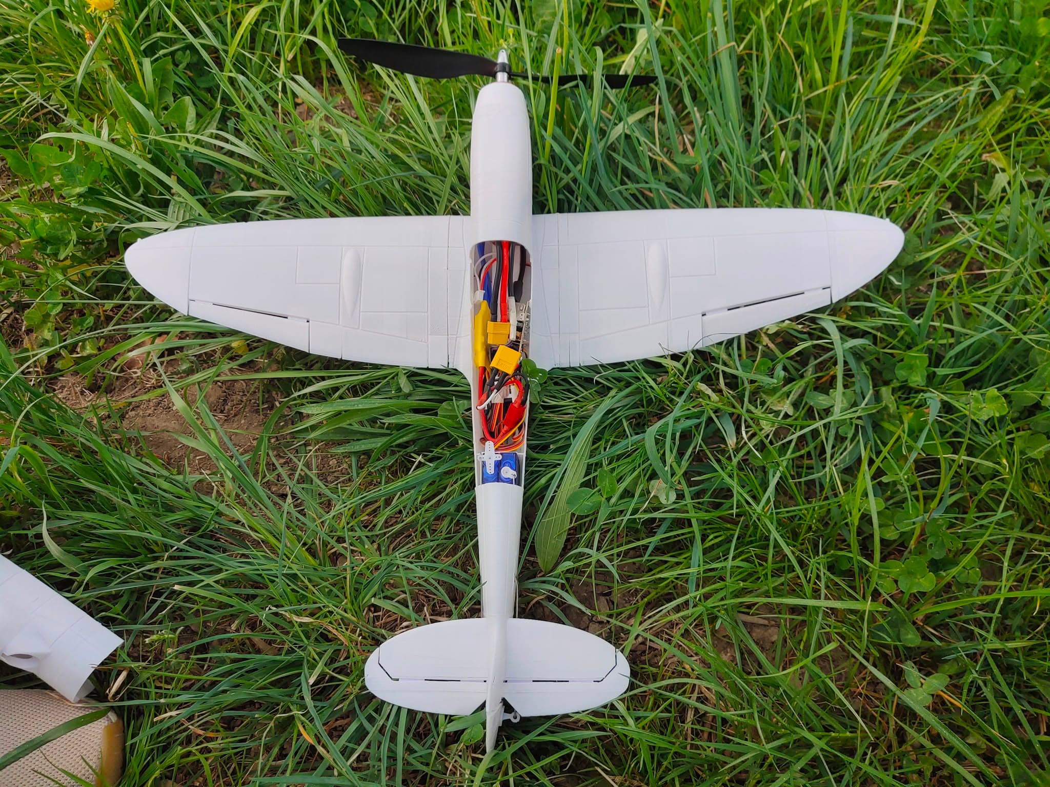 Top view of the unpainted 3D-printed Spitfire lying in the grass before its maiden flight, with the electronics bay open showing ESC, wiring and servos